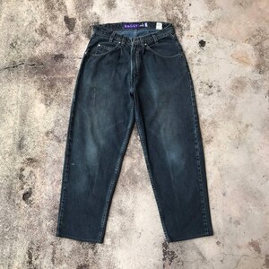 May include: Dark wash denim baggy jeans. The jeans have a relaxed fit with a high waist and a faded appearance. The word "BAGGY" is visible on a purple tag. The jeans are laid flat on a concrete surface.