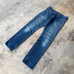 Puede incluir: Un par de vaqueros de mezclilla azul con un lavado descolorido. Los vaqueros est&aacute;n colocados planos sobre una superficie de hormig&oacute;n, mostrando el corte recto y el dise&ntilde;o cl&aacute;sico de cinco bolsillos. La mezclilla tiene marcas de desgaste sutiles.