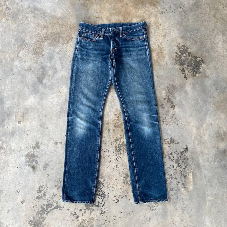 Puede incluir: Un par de vaqueros de mezclilla azul con bragueta de botones y dise&ntilde;o cl&aacute;sico de cinco bolsillos. Los vaqueros tienen un lavado descolorido y sutiles marcas de desgaste, d&aacute;ndoles un aspecto vintage. Los vaqueros est&aacute;n colocados sobre una superficie de hormig&oacute;n.