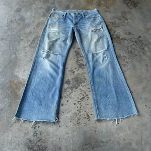 May include: Light blue denim jeans with distressed details, including rips at the knees and a raw hem. The jeans have a wide, flared leg design. The garment is laid flat on a concrete surface.