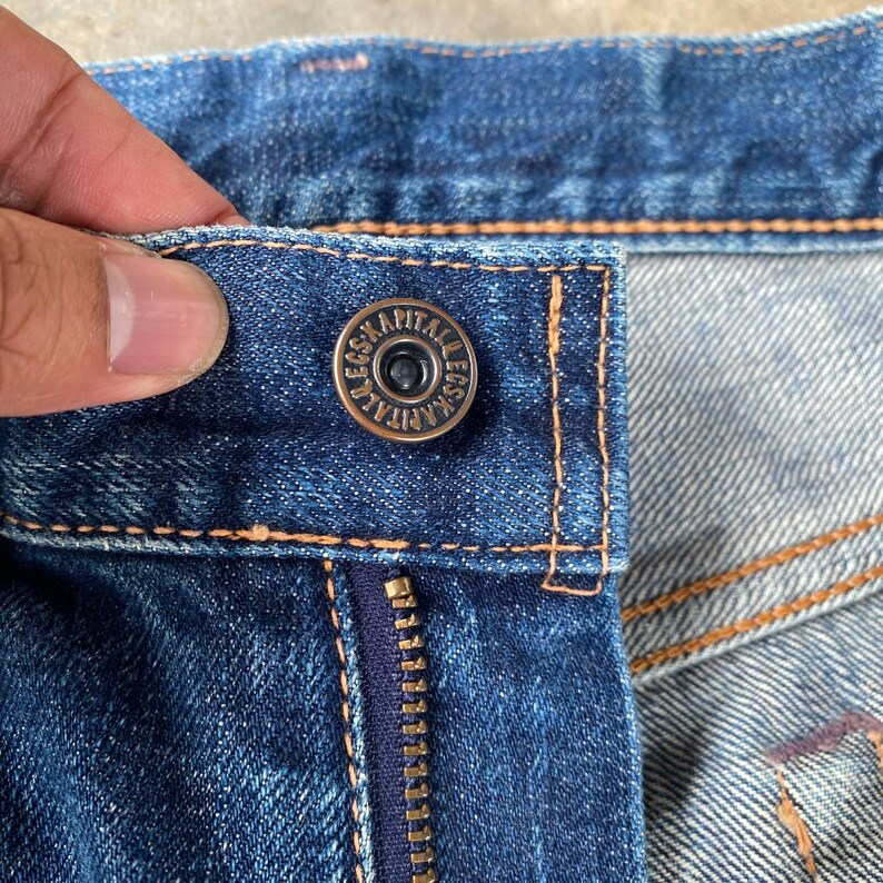 Puede incluir: Primer plano de unos vaqueros azules con un bot&oacute;n met&aacute;lico y una cremallera. El bot&oacute;n tiene el texto "LEGS KAPITALERS" en el borde. Los vaqueros tienen costuras naranjas.
