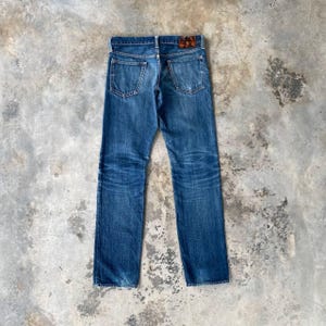 Puede incluir: Vaqueros azules de mezclilla con un dise&ntilde;o cl&aacute;sico de cinco bolsillos. Los vaqueros tienen un lavado descolorido y marcas de desgaste visibles, con un parche de cuero marr&oacute;n en la parte trasera. Los vaqueros est&aacute;n colocados planos sobre una superficie de hormig&oacute;n.