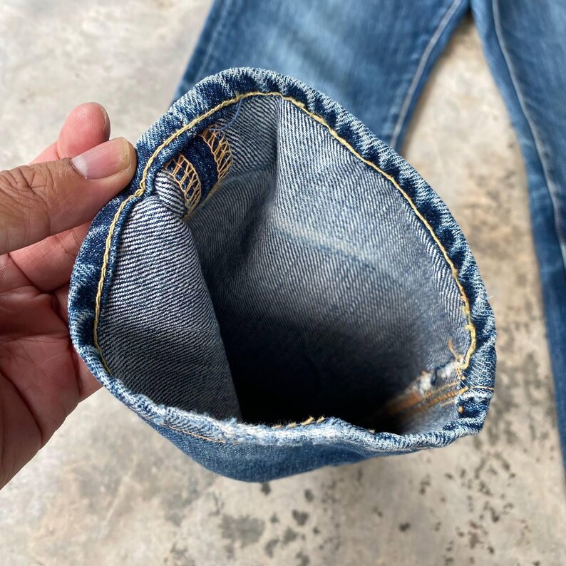 Puede incluir: Primer plano del dobladillo de unos vaqueros azules. Los vaqueros est&aacute;n doblados, revelando la costura interior y las costuras amarillas. La mezclilla tiene un aspecto descolorido y desgastado.