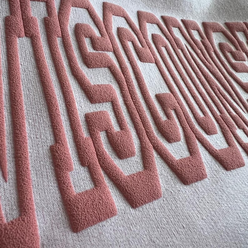 May include: A light-colored textile featuring raised, textured embroidery in a dusty rose color. The embroidery forms a series of interconnected, maze-like geometric lines and shapes that create a three-dimensional, tactile surface pattern.