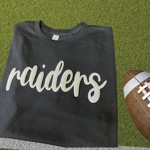 May include: A black short-sleeved t-shirt featuring the word raiders printed in a white, glittery, cursive script font across the chest. The shirt has a crew neckline and is displayed alongside a brown leather American football with white laces.