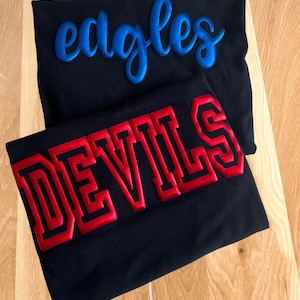 May include: Two black shirts with embroidered text. One shirt features the word "Eagles" in blue script, and the other displays "DEVILS" in red block letters with a red outline. The shirts are displayed on a wooden surface.