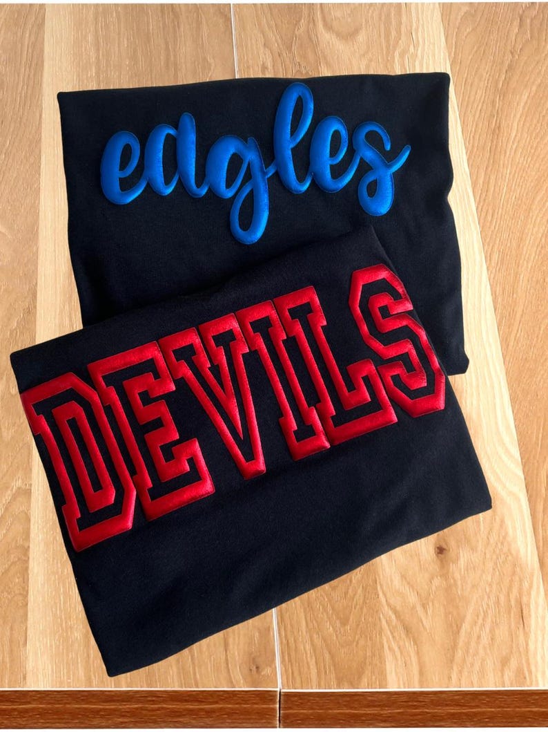 May include: Two black t-shirts stacked on top of each other. The top shirt features the word "eagles" in a blue, raised, cursive font. The bottom shirt features the word "DEVILS" in a red, raised, block-style font with a black outline.