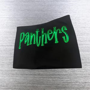 May include: A black t-shirt featuring the word panthers written in a stylized, glossy, bright green font across the chest. The lettering has a raised, three-dimensional appearance with a smooth, reflective surface texture.