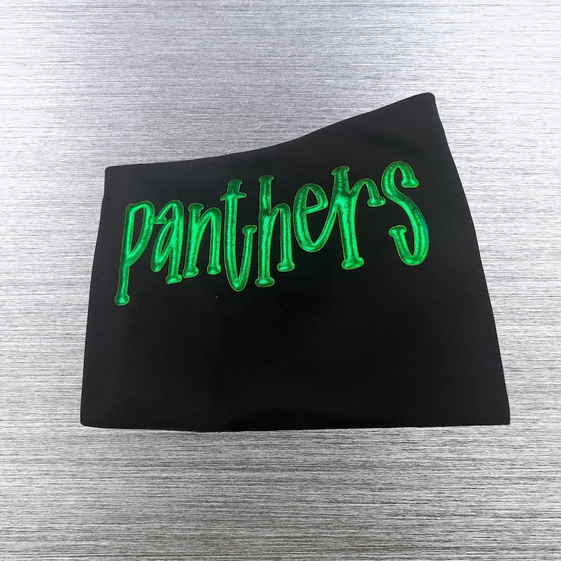 May include: A black t-shirt featuring the word panthers written in a stylized, glossy, bright green font across the chest. The lettering has a raised, three-dimensional appearance with a smooth, reflective surface texture.