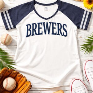May include: White baseball-style t-shirt with navy blue sleeves and the word "BREWERS" in navy blue across the chest. The sleeves have white stripes. Baseball and glove are in the foreground.