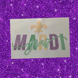May include: A white fabric with a purple, green, and gold rhinestone design that reads "Mardi Gras" with a fleur-de-lis above the text.