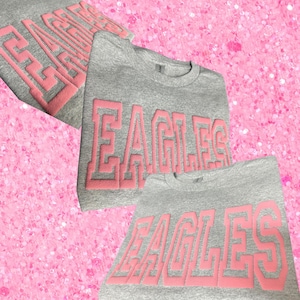 May include: A heather gray crewneck sweatshirt featuring the word EAGLES in large, raised, light pink block lettering across the chest. The garment has a ribbed collar and long sleeves, with the design repeated in a layered, collage-style layout.