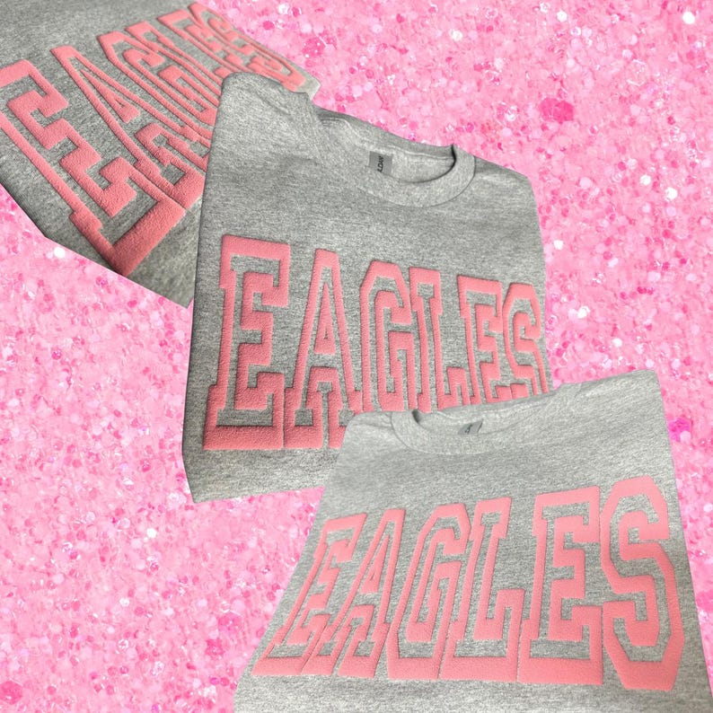 May include: A heather gray crewneck sweatshirt featuring the word EAGLES in large, raised, light pink block lettering across the chest. The garment has a ribbed collar and long sleeves, with the design repeated in a layered, collage-style layout.