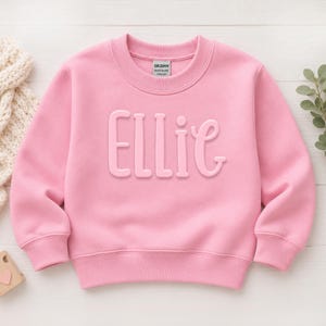 May include: A light pink long-sleeve sweatshirt featuring the name Ellie in raised, matching pink lettering across the chest. The garment has a crew neckline, ribbed cuffs, and a ribbed hem, with a Gildan brand label visible at the inner neck.
