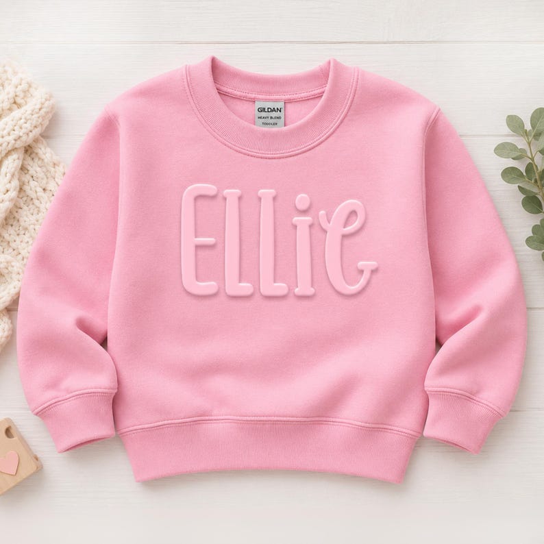 May include: A light pink long-sleeve sweatshirt featuring the name Ellie in raised, matching pink lettering across the chest. The garment has a crew neckline, ribbed cuffs, and a ribbed hem, with a Gildan brand label visible at the inner neck.