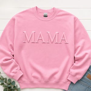 May include: A light pink crewneck sweatshirt featuring the word MAMA embossed in a matching pink tone across the chest. The garment has long sleeves, a ribbed collar, and a Gildan brand label visible at the inner neckline, paired with blue denim.