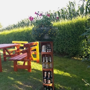 May include: A wooden beer bottle holder with multiple shelves, filled with various beer bottles. The holder is in a garden setting with a picnic table and a flower pot.