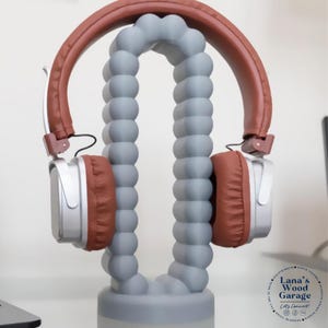 May include: A pair of headphones rests on a gray, decorative stand. The headphones have brown earcups and headband, with silver accents. The stand is a light gray color and has a unique, curved design. The stand is labeled "Lana's Wood Garage".