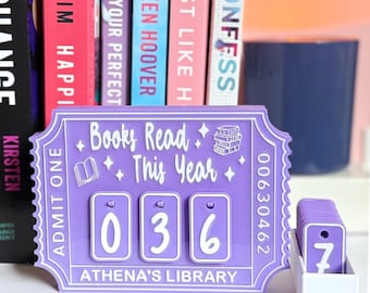 May include: A purple "Books Read This Year" counter with the number "036" displayed. The counter has a ticket design with the text "ATHENA'S LIBRARY" and "ADMIT ONE". A small box with the number "7" is next to the counter. Books are in the background.