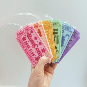 May include: A hand holds a fan of six colorful, ticket-shaped tags. The tags are pink, peach, orange, green, blue, and purple. The pink tag reads "RomCom Book Club" and "Lifetime Membership". Each tag has a clear plastic loop.