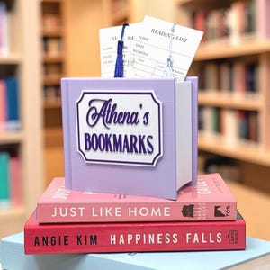 May include: A lavender-colored book-shaped bookmark holder labeled "Athena's BOOKMARKS" with reading list cards and a blue tassel. The holder sits atop a stack of books with titles like "JUST LIKE HOME" and "HAPPINESS FALLS".