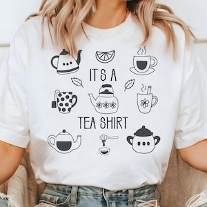 May include: White t-shirt with a black graphic design featuring teapots, teacups, and the text "It's a Tea Shirt." The design includes a lemon slice, leaves, and a honey dipper. The shirt is a casual, everyday wear item.
