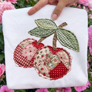 May include: White sweatshirt featuring a patchwork cherry design. The cherries are made of red and pink patterned fabric, with green gingham and polka dot leaves. The design is hand-stitched.