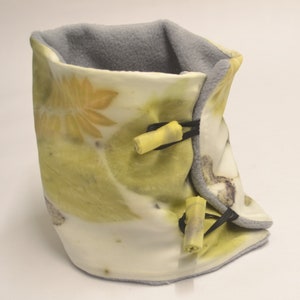 May include: A neck warmer featuring a cream-colored exterior with botanical leaf prints in shades of yellow and green. The garment is lined with soft grey fleece and includes two yellow toggle closures secured by black elastic loops on the front side.