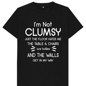 Peut inclure: T-shirt noir avec texte blanc : « I'm Not CLUMSY, JUST THE FLOOR HATES ME, THE TABLE & CHAIRS are bullies, AND THE WALLS GET IN MY WAY ». Le t-shirt a un col rond et des manches courtes.