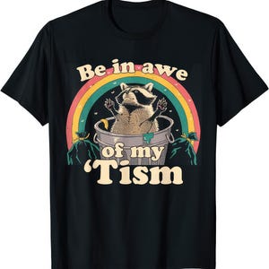 May include: Black t-shirt with a raccoon design inside a bin, with a rainbow arch. The text reads "Be in awe of my 'Tism". The raccoon is tan and black, with its paws raised. The design has a vintage, retro style.