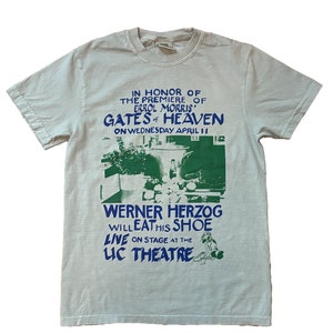 May include: A light blue t-shirt with a green and blue print advertising a Werner Herzog event. The print includes the text "In honor of the premiere of Errol Morris' Gates of Heaven on Wednesday April 11" and "Werner Herzog will eat his shoe live on stage at the Luc Theatre."