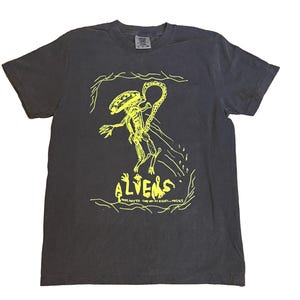 May include: Dark gray t-shirt featuring a neon yellow illustration of an alien with a long tail. The word "ALIENS" is written in a stylized font below the alien. The shirt has a crew neck and short sleeves.