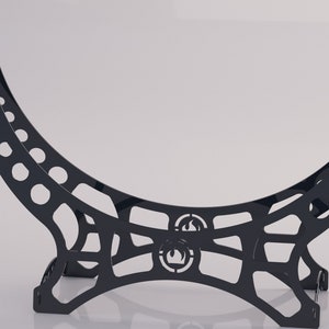Puede incluir: Soporte de metal negro con un diseño curvo y una abertura circular. El soporte tiene un patrón decorativo con un logotipo de llama.