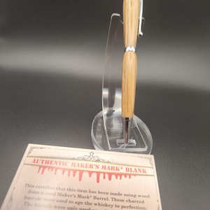May include: A wooden pen with silver accents and a silver tip, displayed on a clear stand. The pen is made from wood from a Maker's Mark barrel. A card below the pen reads "AUTHENTIC MAKER'S MARK BLANK".