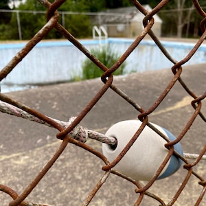 May include: A rusty chain link fence with a white and blue buoy attached to it. The buoy is hanging from a rope and is partially obscured by the fence. The fence is in front of an empty swimming pool.