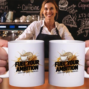 Peut inclure: Deux mugs en céramique blanche avec le texte "FUEL YOUR AMBITION" et un dessin d'une tasse à café et d'un cerveau. Les tasses sont tenues par des mains devant une femme souriante dans un cadre de café. Les tasses ont aussi le texte "IDEAS DON'T BREW THEMSELVES."