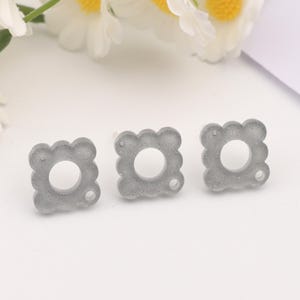 Pode incluir: Três peças em branco para brincos quadrados de resina cinza com um furo redondo no centro. Cada peça em branco tem uma superfície texturizada.