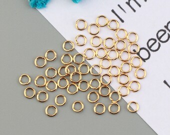 May include: A large quantity of gold jump rings, scattered on a white and gray surface. The rings are approximately 6mm in diameter.