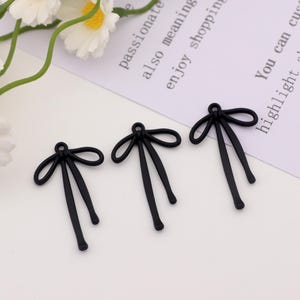 May include: Three black bow-shaped charms with long, dangling ties. The charms are arranged on a white surface, with a sprig of white flowers and text that reads "passionate" and "enjoy shopping" in the background. These charms are ideal for jewelry making.
