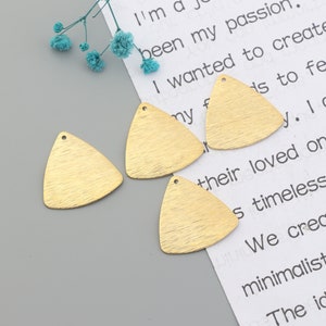 May include: Four gold-colored, triangle-shaped metal blanks with a brushed texture. Each blank has a small hole at the top. The blanks are on a gray surface with a piece of paper with text and some blue flowers.