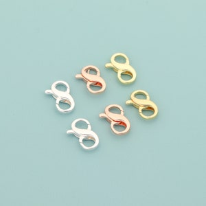 Könnte beinhalten: Sechs Metall-Hummerverschlüsse in Silber, Roségold und Goldtönen sind auf einem hellblauen Hintergrund angeordnet. Die Verschlüsse sind geöffnet und zeigen ihr Haken- und Schlaufendesign, bereit für die Schmuckherstellung.