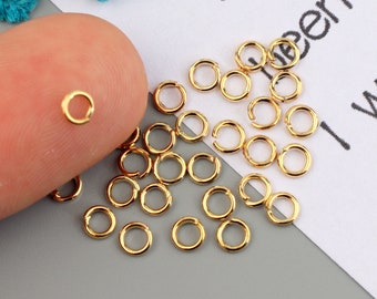 May include: A collection of small, gold-colored jump rings, some open and some closed, scattered on a gray surface. One ring is held by a finger. These jewelry findings are used for connecting jewelry components.