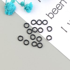 May include: A close-up of a group of black jump rings. The rings are scattered on a white surface with a gray background.