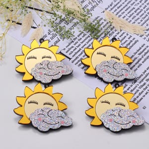 May include: Four acrylic sun and cloud charms. Each charm features a yellow sun with closed eyes and a silver glitter cloud with a pink smiling face. These charms are ideal for jewelry making or craft projects.
