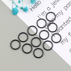 Puede incluir: Primer plano de diez anillos de salto de metal negro. Los anillos están dispuestos en círculo sobre una superficie blanca con texto.