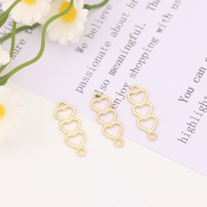 Puede incluir: Tres abalorios de metal de color dorado en forma de corazón, cada corazón es ligeramente más pequeño que el de arriba, los abalorios están conectados por un bucle en la parte superior del corazón más grande.