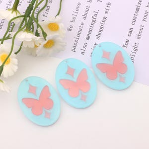 Puede incluir: Tres abalorios ovalados de acrílico azul claro con un diseño de mariposa rosa y un agujero plateado en la parte superior. Cada abalorio tiene un diseño de diamante brillante alrededor de la mariposa.