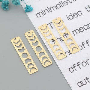 May include: Four gold-colored rectangular charms with a moon phase design. Each charm has a small hole at the top for stringing. The charms are arranged on a gray surface with a piece of paper with text.