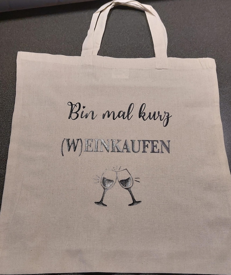 Könnte beinhalten: Eine weiße Stofftasche mit schwarzem Sticktext, der "Bin mal kurz (W)EINKAUFEN" lautet, und einer Grafik von zwei sich anstoßenden Weingläsern.