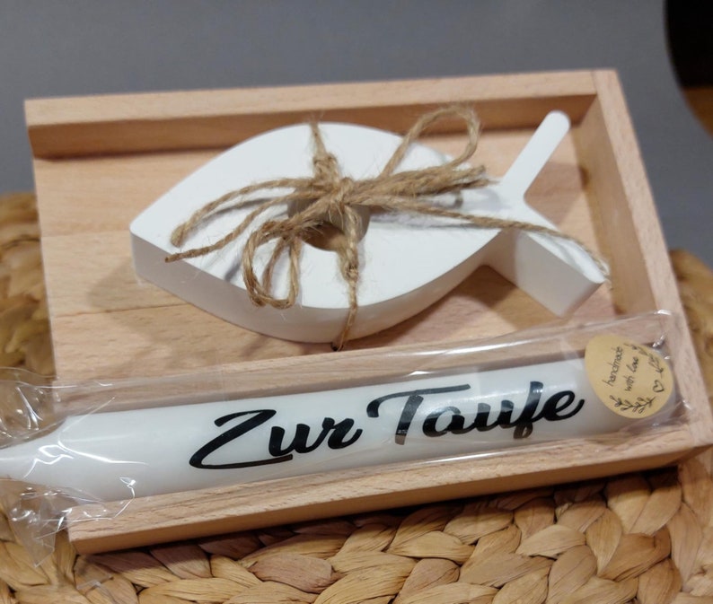 Könnte beinhalten: Eine Holzkiste mit einem weißen Holzfisch und einer weißen Kerze. Die Kerze ist in Plastik verpackt und trägt den Text "Zur Taufe". Es gibt ein kleines braunes Etikett mit dem Text "handgefertigt mit Liebe" darauf.